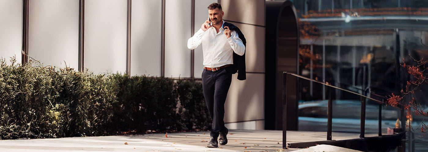 Un homme entre 25 et 40 ans marche avec un veston sur l'épaule, une chemise blanche uni et un pantalon propre
