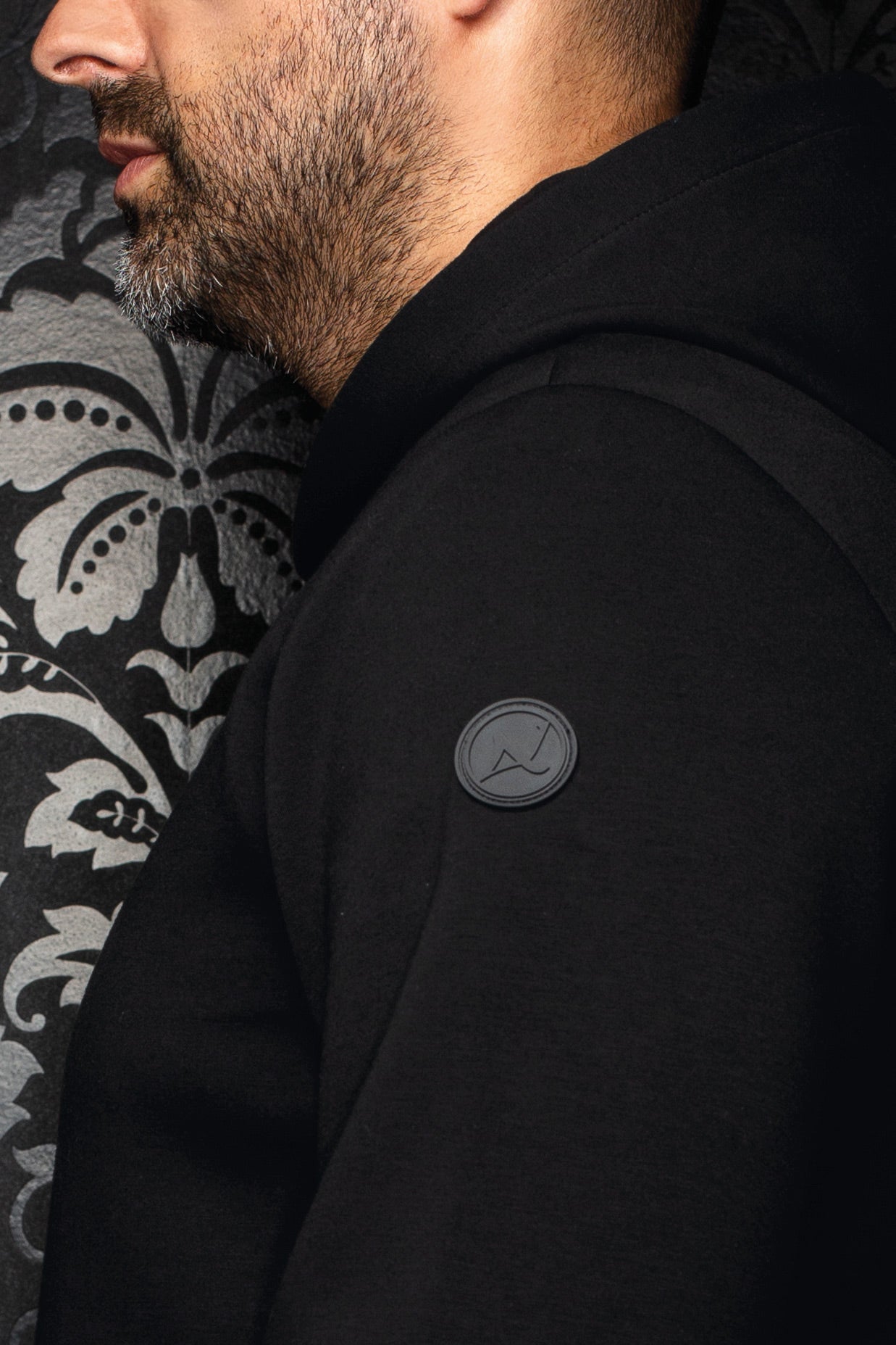 Côté d’une veste sport noire à capuchon avec couture raglan et écusson ton sur ton pour homme par Au Noir, en gros plan
