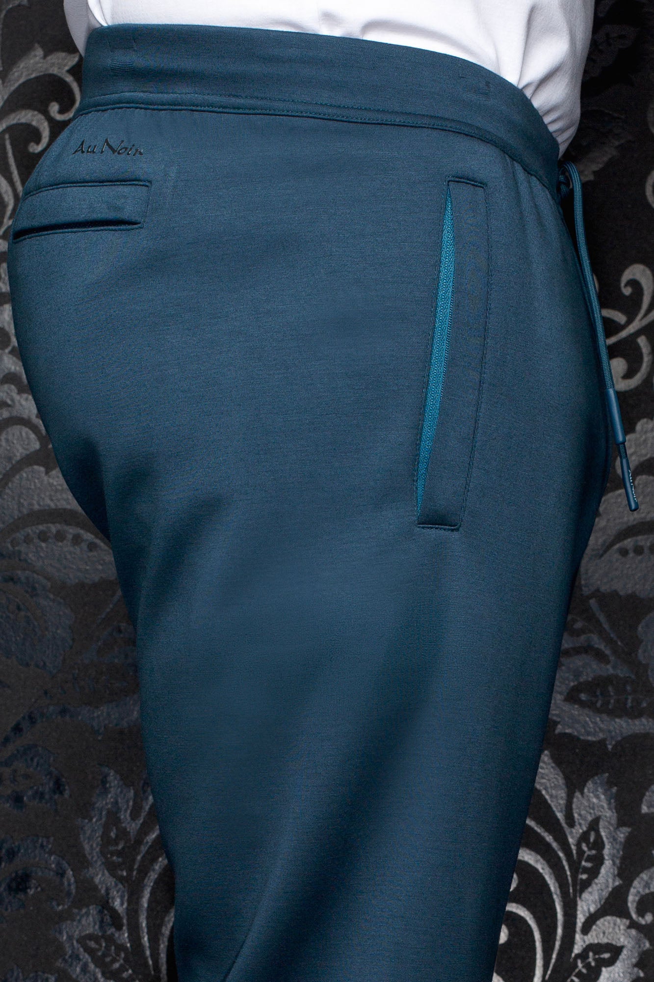 Men's teal jogging pants with elastic waistband, back welt pocket and Au Noir logo, close-up