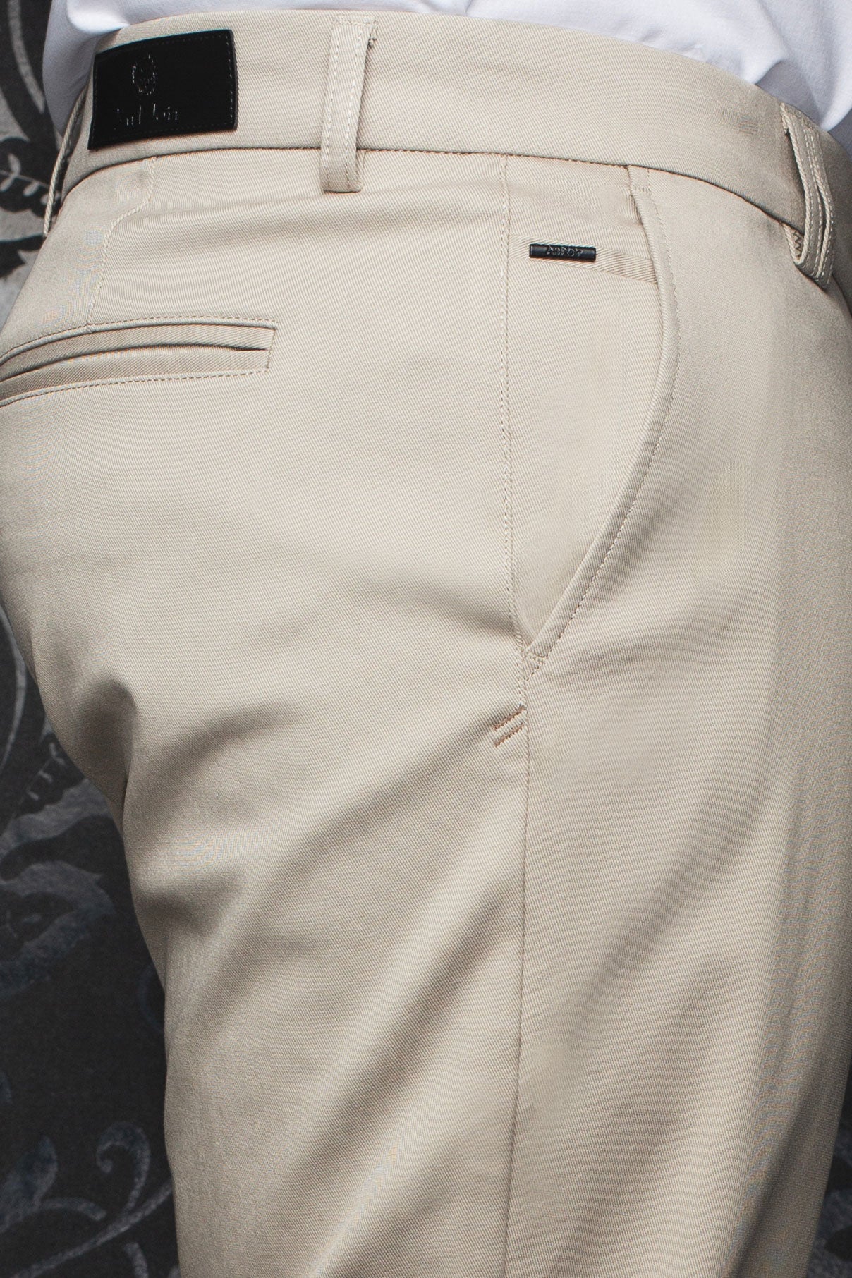 Détail rapproché du pantalon chino beige avec poche passepoilée, passants et écusson noir pour homme par Au Noir